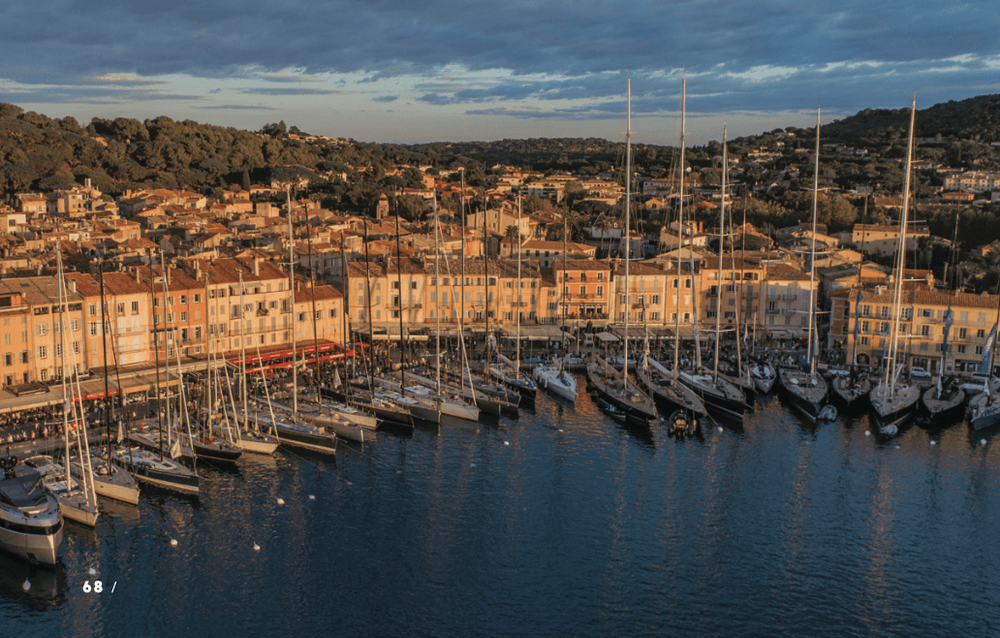 Sailboats fill a Mediterranean harbor, fronting a colorful town under a warm evening sky.