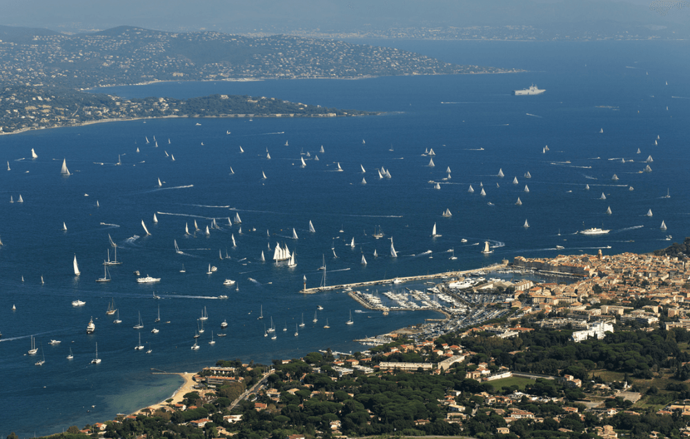 Aerial view of a busy bay with numerous white sailboats and yachts on blue water.