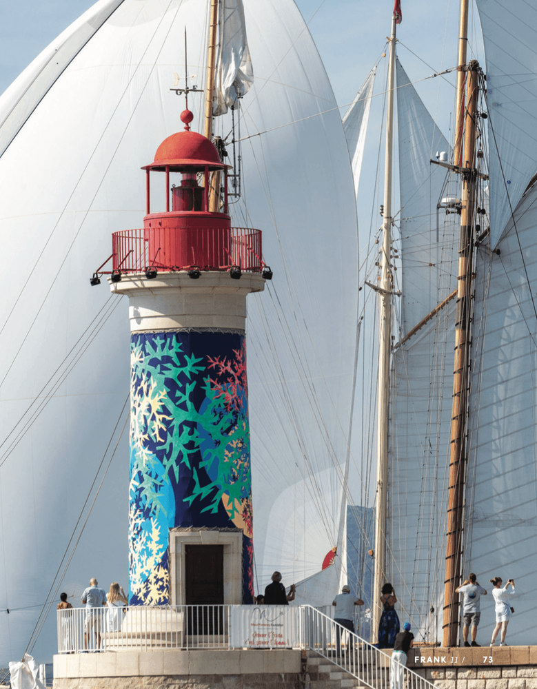 Red and white lighthouse with vibrant artistic base, towering over large white sailboat sails.