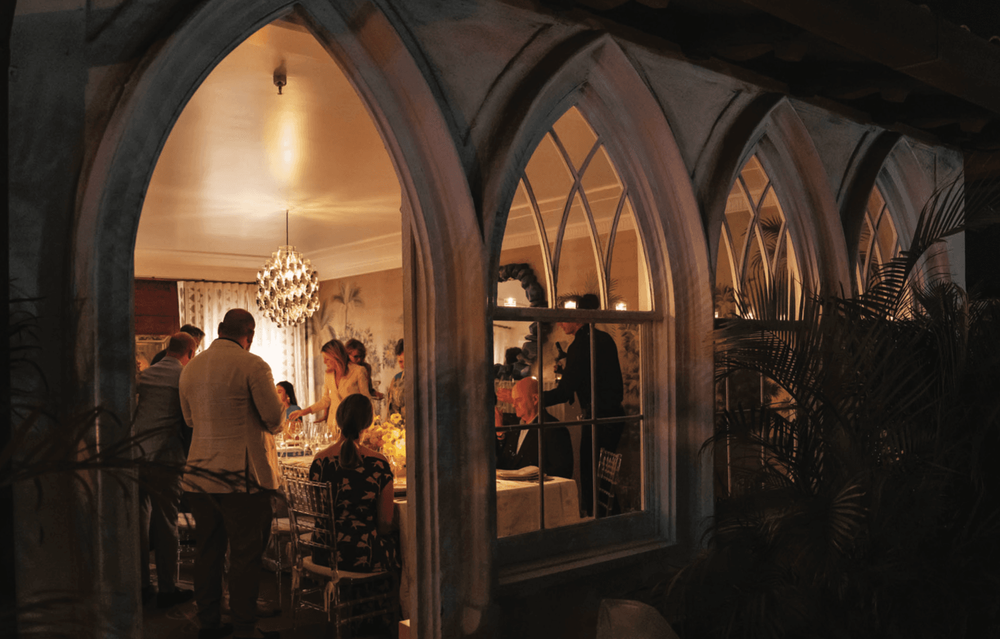 People gather inside an elegantly illuminated room with distinctive arched windows at night.