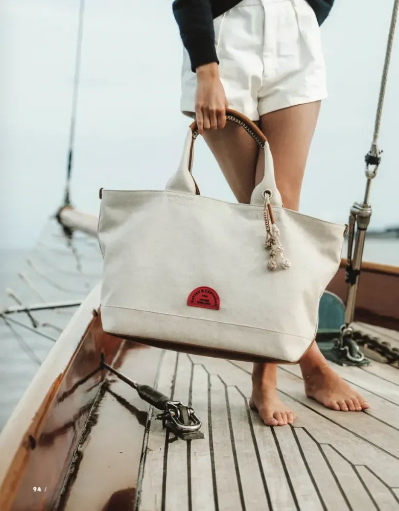 Barefoot person on wooden boat deck holding off-white canvas tote bag with red design.