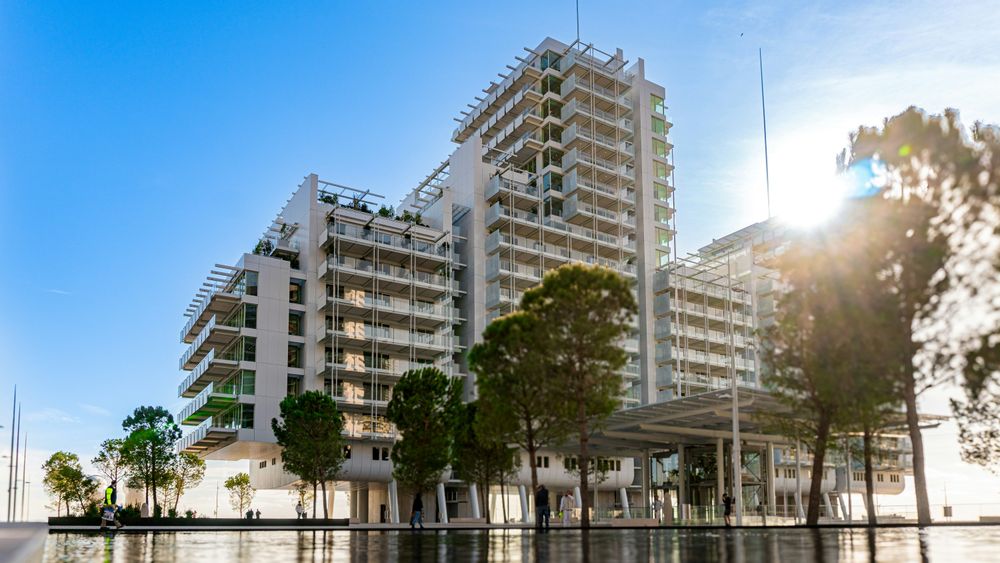 Modern white building with balconies and trees under a bright blue sky on a sunny day.