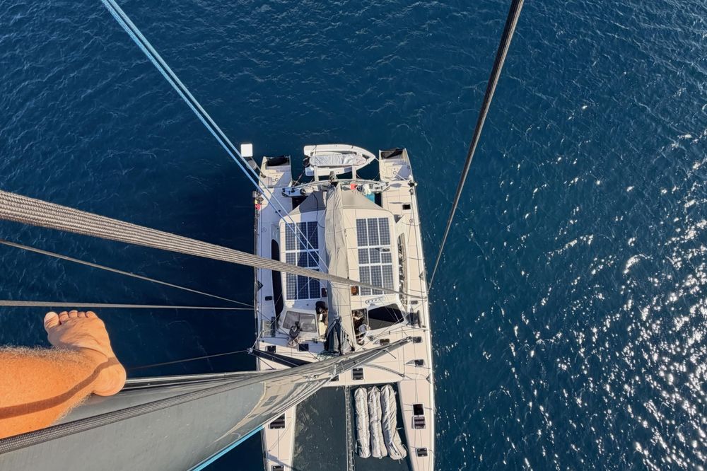 Overhead view from mast of white catamaran with solar panels on calm blue water.