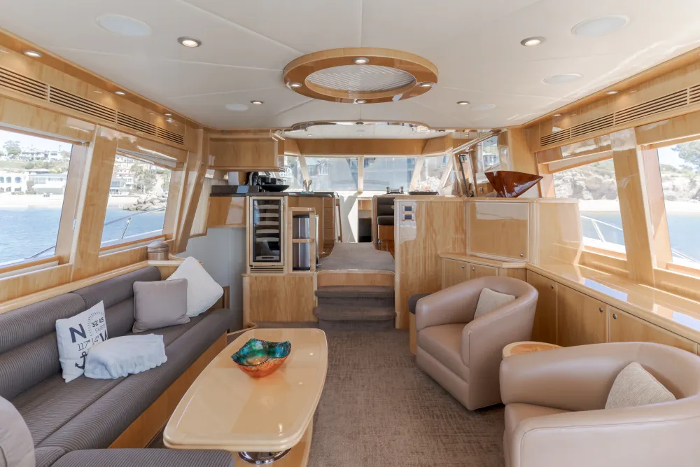 Luxurious yacht interior with light wood, grey sofa, beige chairs, and large windows.