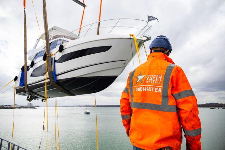 A man in an orange YACHT LIFTMASTER jacket watches a white yacht being craned over water.