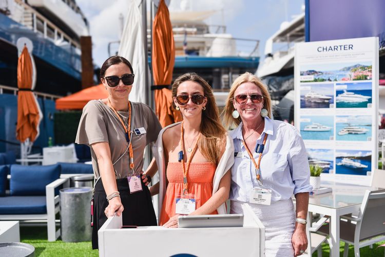 Three women smiling at a luxury yacht charter exhibition outdoors.
