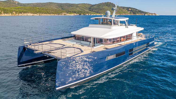 Large blue catamaran yacht glides across clear blue water with green hills in background.