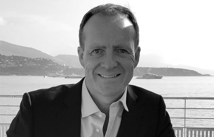 Smiling man in a suit jacket with a scenic outdoor background of water and mountains.