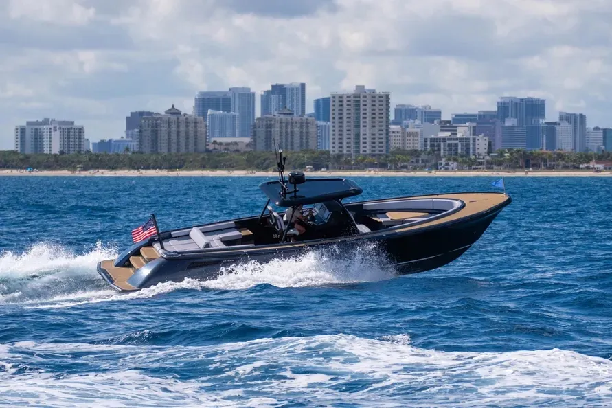 Sleek dark speedboat racing across blue ocean water with a distant city skyline.