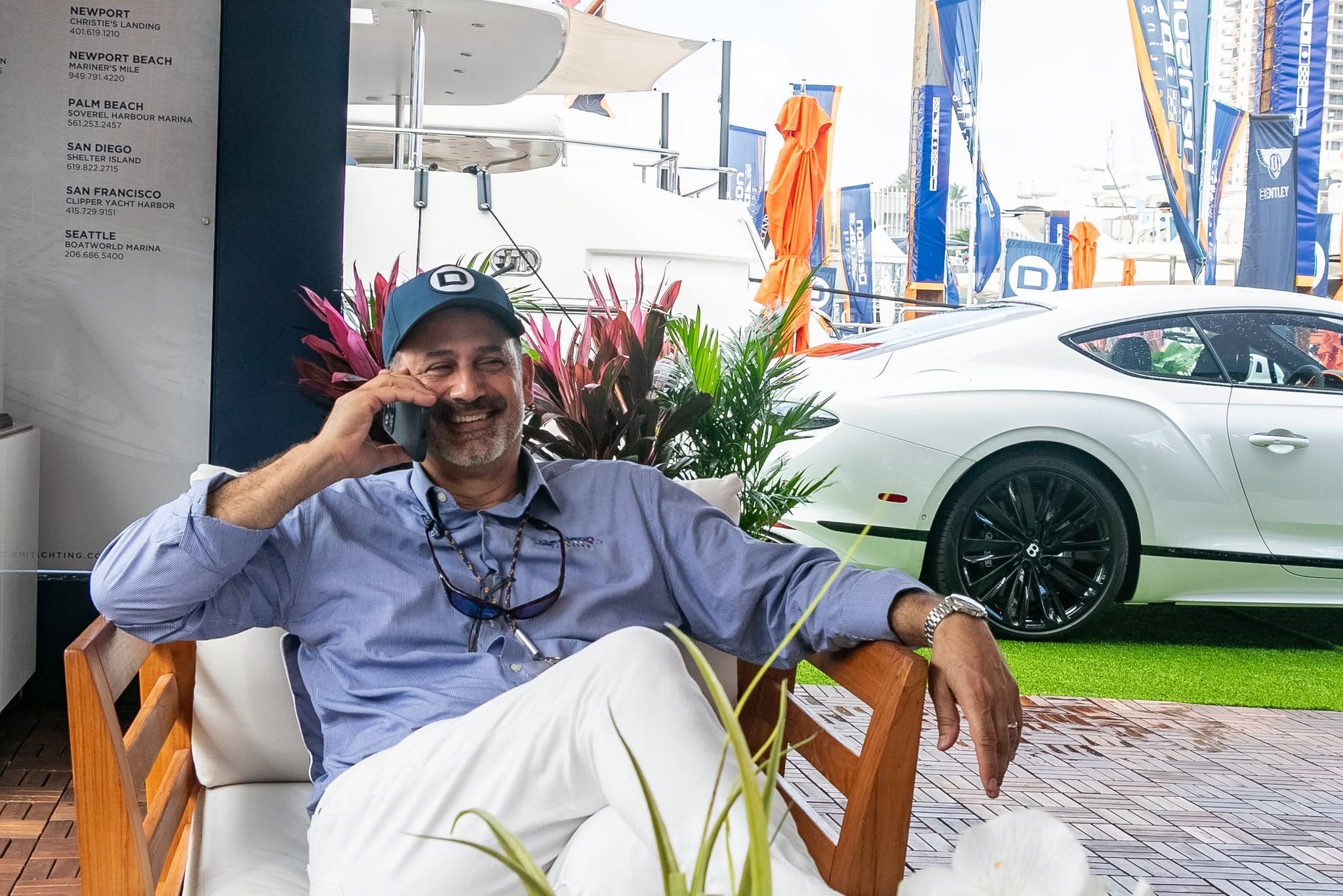 Smiling man in blue shirt and cap talks on phone, luxury car and boats visible.