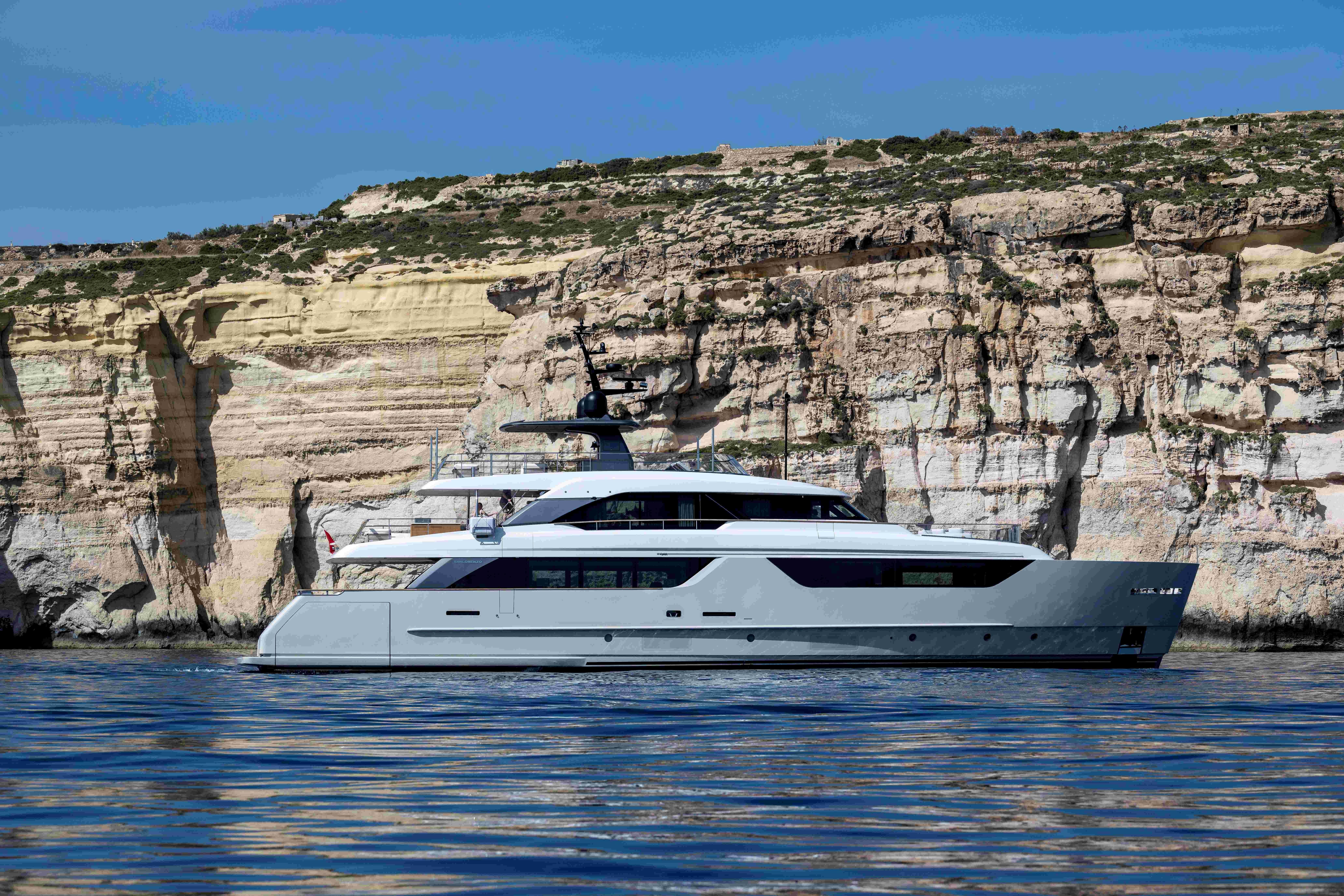 Modern grey yacht cruises on blue water beside a towering rocky cliff.