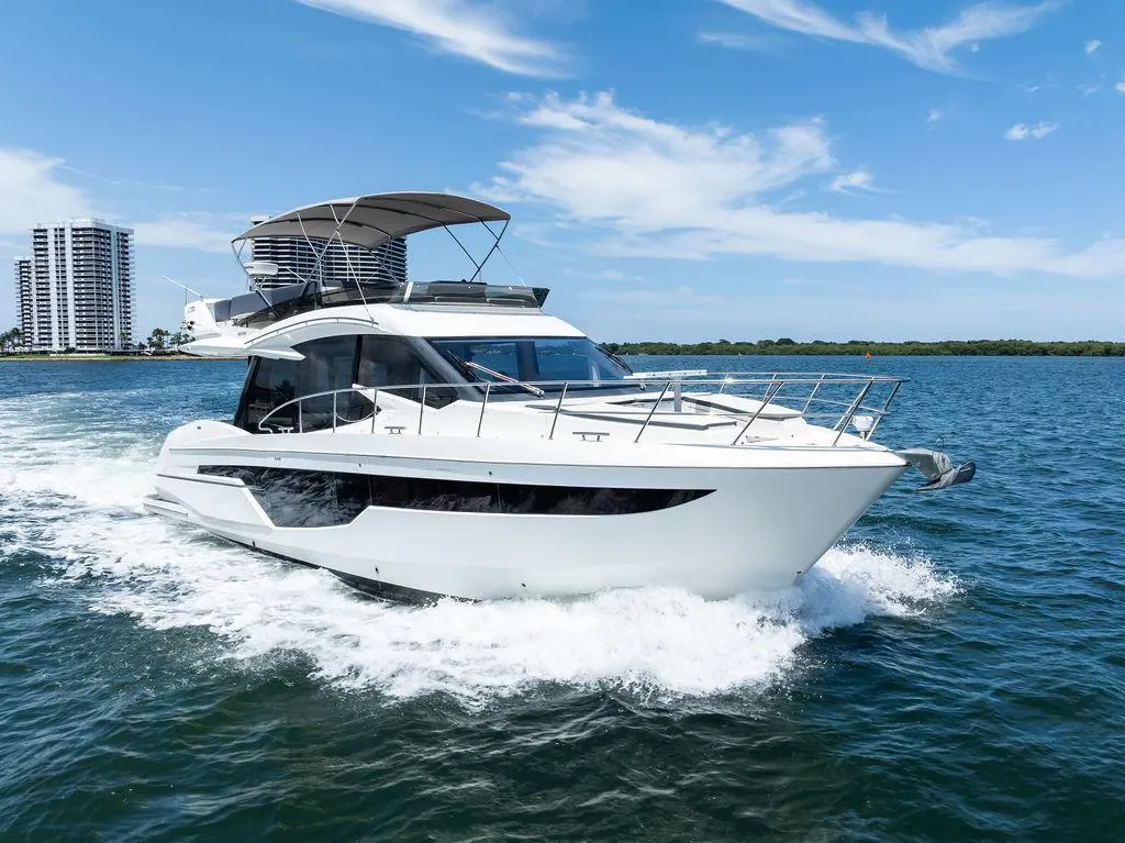 White luxury yacht cruising on blue water, leaving a wake, with a city skyline visible.
