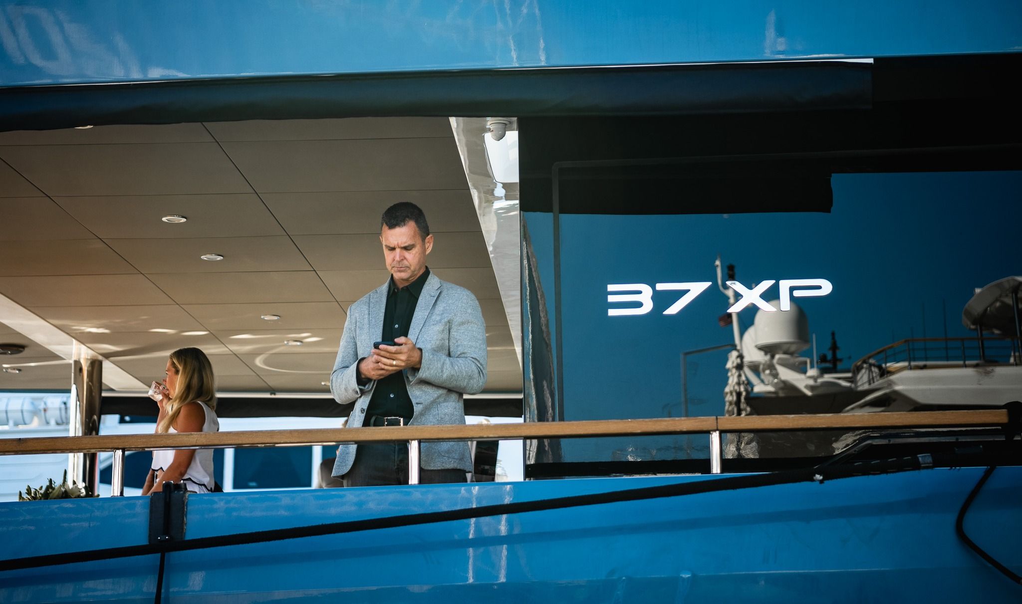 Man looking at phone on a blue boat with '37 XP' text, woman stands behind him.