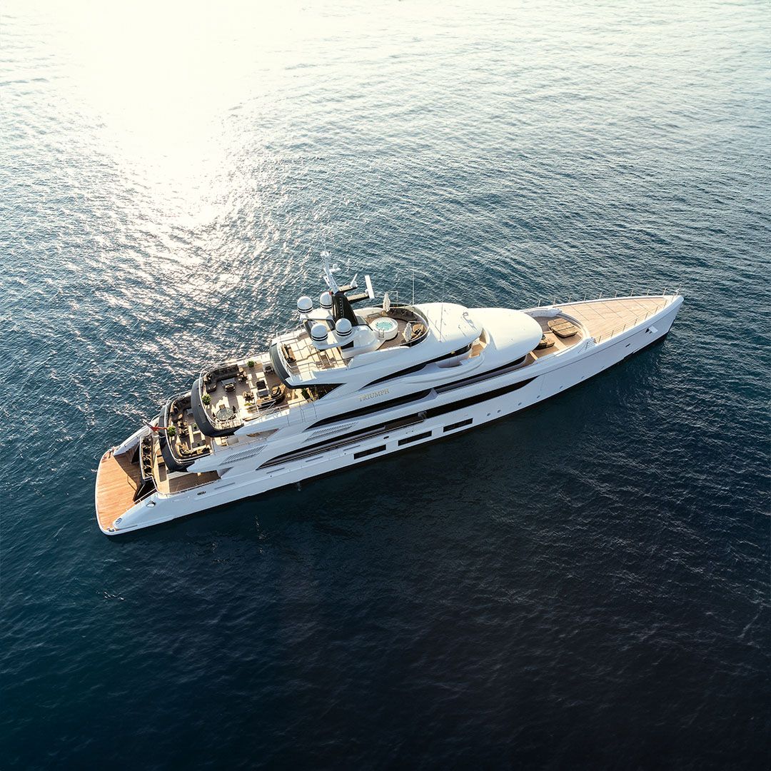 A large, sleek white superyacht with multiple decks sails across calm blue ocean waters.