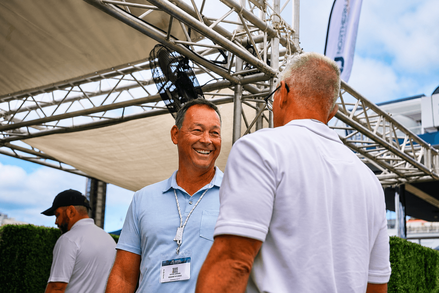 Two men smiling and talking under a tent at an outdoor event.