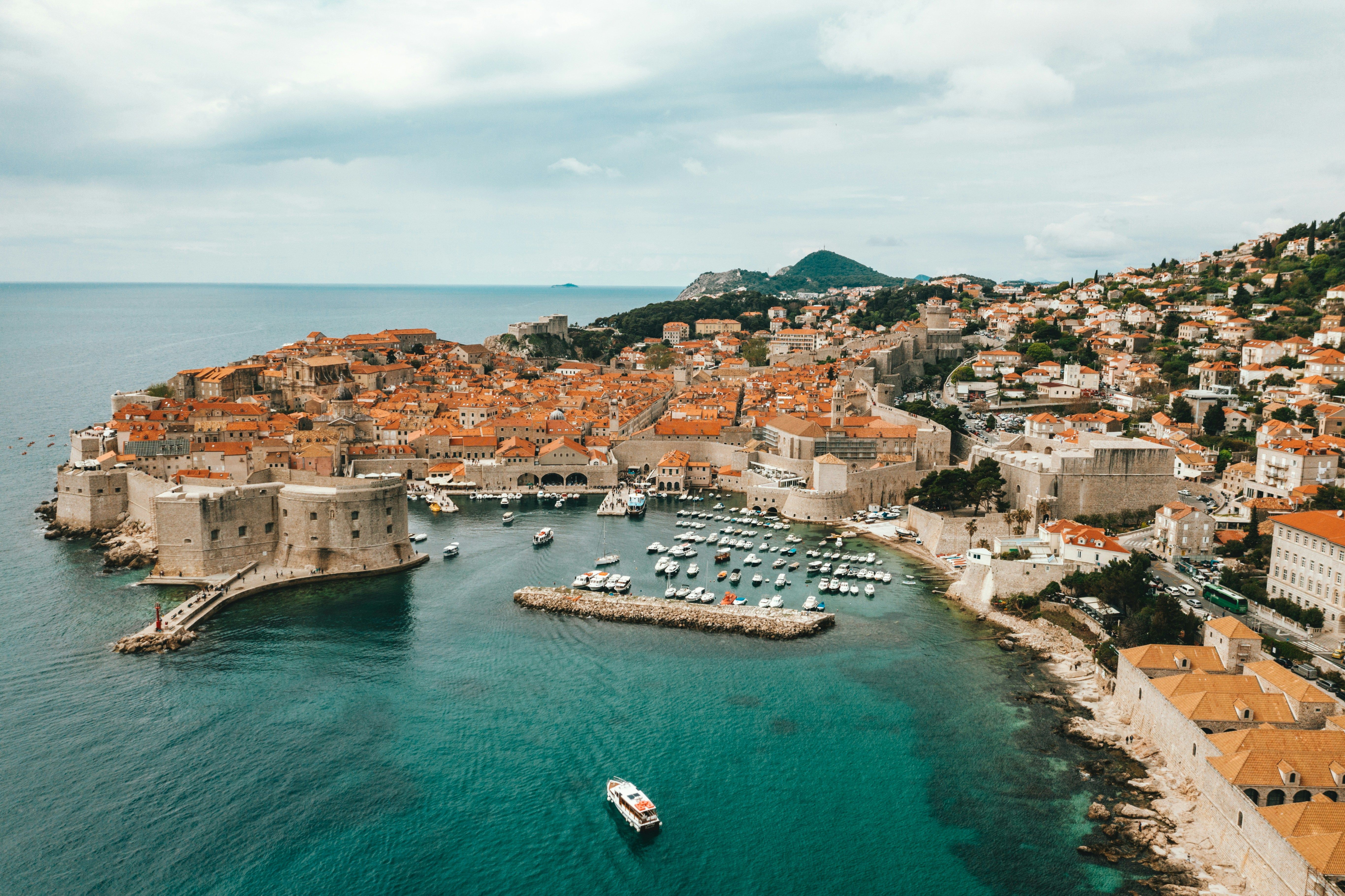 Aerial view of ancient coastal city with red roofs, walled fortress, harbor, and boats.