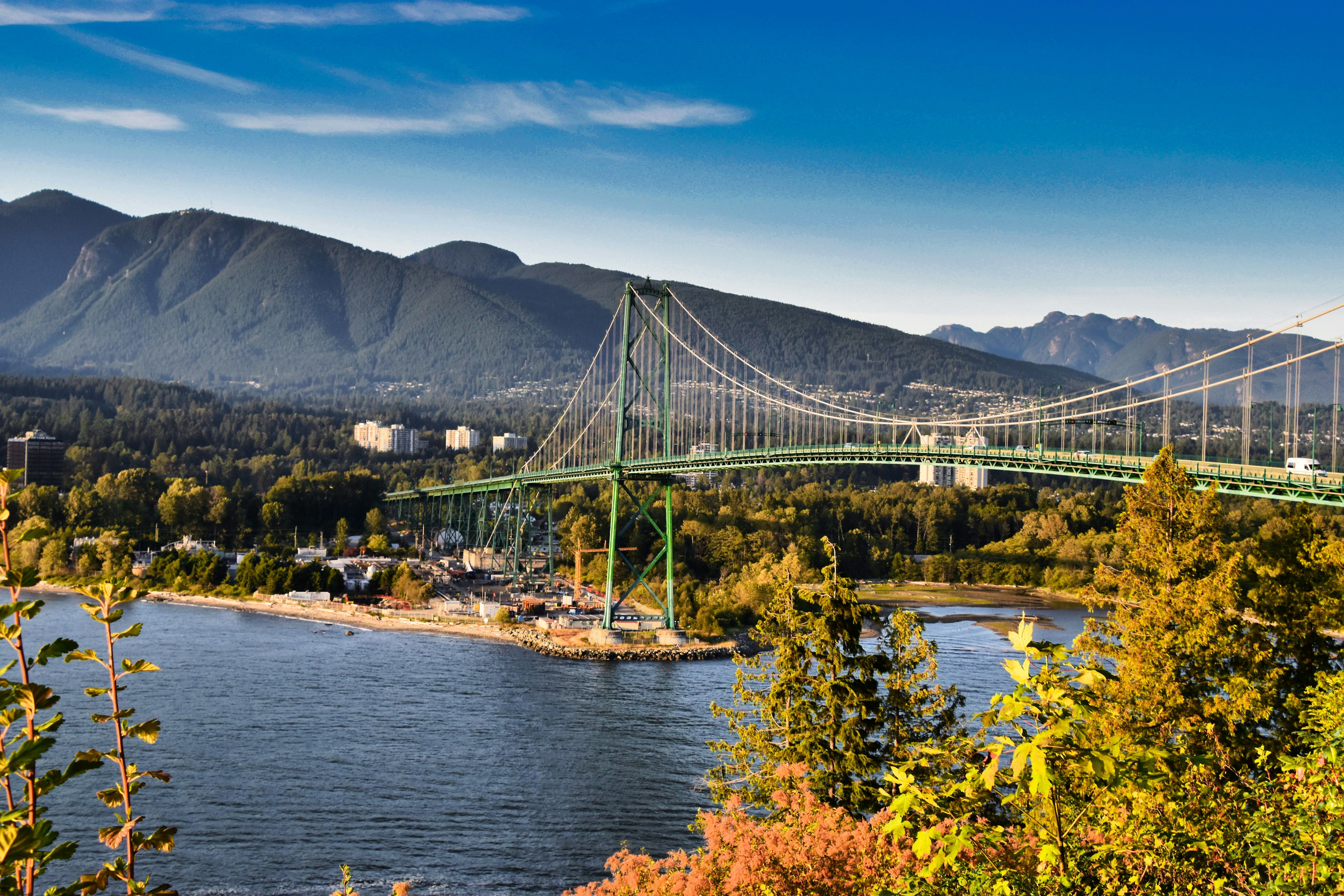 Green suspension bridge spanning blue water with forested mountains and autumn foliage.